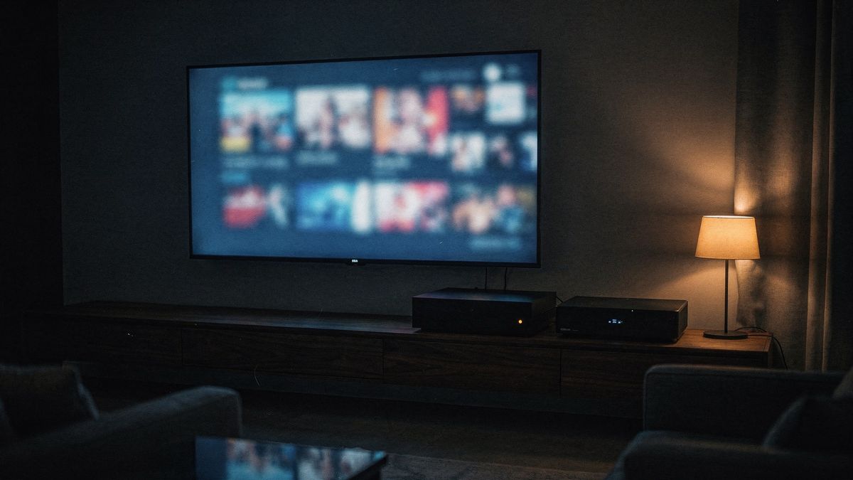 A living room at night. A television shows a wall of film thumbnails; on the shelf below, a small black server box with one amber light.