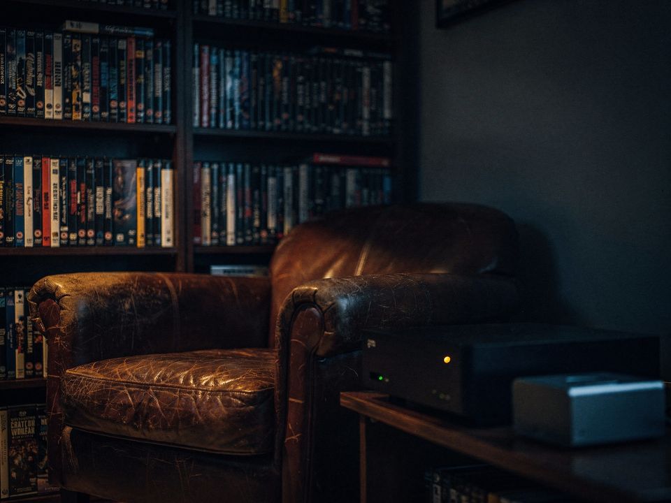 A dim room corner: shelves packed with DVD and Blu-ray cases, a worn leather armchair, and on a low shelf a small black box with one amber light.